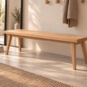 Könnte beinhalten: Eine rechteckige Holzbank mit hellbrauner Oberfläche, bestehend aus einer flachen Sitzfläche und vier konisch zulaufenden Beinen, die nach außen gerichtet sind. Die Bank ist aus Naturholz mit sichtbarer Maserung gefertigt und stabil.