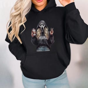 May include: Black hoodie featuring a graphic of a Scream mask with blonde hair, holding a coffee cup and making a peace sign. The word "MAMA" is printed on the hoodie. The hoodie has a front pocket and a hood.