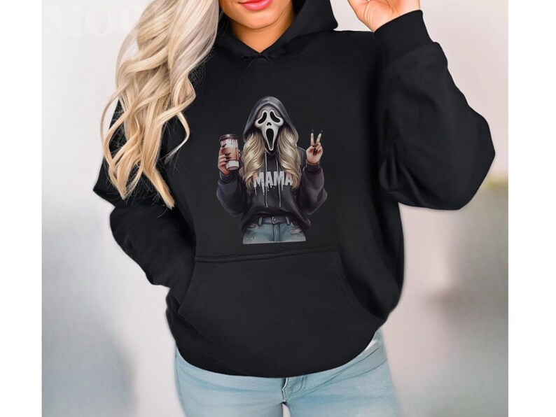 May include: Black hoodie featuring a graphic of a Scream mask with blonde hair, holding a coffee cup and making a peace sign. The word "MAMA" is printed on the hoodie. The hoodie has a front pocket and a hood.