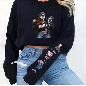 May include: Black sweatshirt featuring a graphic of a Scream-masked figure holding a child, with the word "MAMA" on the front. The sleeve displays cartoon characters and names. A Halloween-themed apparel item.