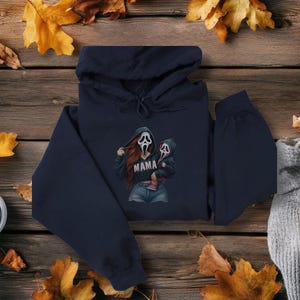 May include: Navy blue hoodie featuring a graphic of a woman and child in Ghostface masks, with the word "MAMA" printed on the front. The image is set against a wooden background with autumn leaves.