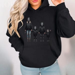May include: Black hoodie featuring a graphic of a family dressed as Ghostface from the Scream movies. The figures are wearing black jackets and holding hands. The adult figure is holding a knife. The hoodie has a front pocket and a hood.