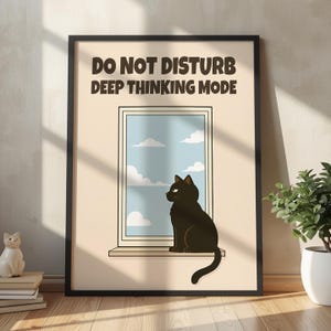 Op de afbeelding: Een ingelijste kunstdruk met een zwarte kat die op een vensterbank zit en uitkijkt over een blauwe lucht met witte wolken. De tekst DO NOT DISTURB DEEP THINKING MODE staat in donkerbruine hoofdletters bovenaan de crèmekleurige achtergrond.