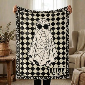Puede incluir: Una manta rectangular con un patrón de cuadros de rombos en blanco y negro y un borde decorativo de volutas. En el centro, una figura de fantasma está cubierta con un diseño de telaraña y lleva gafas de sol redondas negras. El nombre ALICE está impreso.