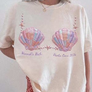 May include: A cream-coloured t-shirt with a seashell design in pastel pink, blue, and orange. The design includes beaded accents and the text "Hannah's Bach Punta Cana 2026" in a script font. The shirt has a relaxed fit.