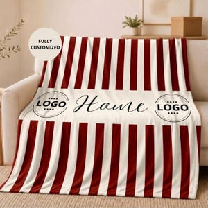 Puede incluir: Una manta con un patrón de rayas verticales de color rojo oscuro y crema. Una banda horizontal central de color crema muestra la palabra Home en letra cursiva negra, flanqueada por dos marcos circulares con el texto LOGO en mayúsculas negras.