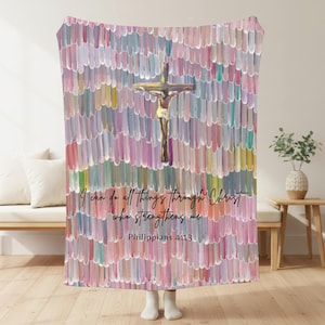 May include: A soft fleece blanket featuring a vertical brushstroke pattern in shades of pink, white, and green. A gold-toned crucifix is centred at the top. The text reads: I can do all things through Christ who strengthens me. Philippians 4:13.
