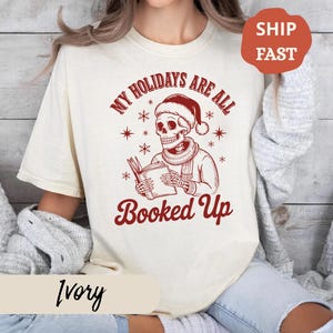 Puede incluir: Camiseta color marfil con un gráfico rojo de un esqueleto con gorro de Papá Noel leyendo un libro. El texto "My Holidays Are All Booked Up" está sobre el esqueleto. Una pegatina roja "Ship Fast" está en la esquina superior derecha.