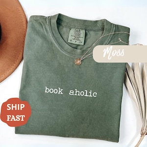 Puede incluir: Una camiseta verde musgo con las palabras "book aholic" impresas en blanco. Un collar dorado con un colgante descansa sobre la camiseta. La camiseta está doblada y tiene una pequeña etiqueta en el cuello. Un sombrero marrón y una planta seca están en el fondo.