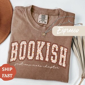 Puede incluir: Camiseta color espresso con la palabra "BOOKISH" en un diseño floral. El texto "just one more chapter" está debajo de la palabra. Un collar plateado con un colgante está sobre la camiseta.
