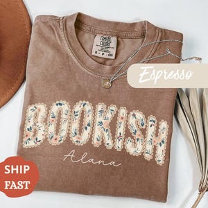 Puede incluir: Camiseta color espresso con la palabra "BOOKISH" en un diseño floral. El nombre "Alana" está escrito debajo. Un collar plateado con un colgante descansa sobre la camiseta. La camiseta tiene una etiqueta Comfort Colors.