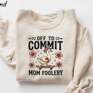 May include: Sand-colored sweatshirt with the text "OFF TO COMMIT MOM FOOLERY". The graphic features a goose wearing a witch hat and holding an apple, riding a skateboard, surrounded by flowers. The sweatshirt has long sleeves and a crew neck.