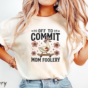 May include: Ivory-colored t-shirt with the text "OFF TO COMMIT MOM FOOLERY." The graphic features a goose wearing a witch hat and riding a skateboard, surrounded by pink flowers. The word "ivory" is printed at the bottom left.