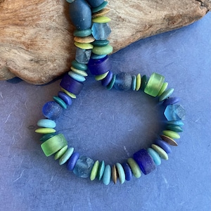 Peut inclure: Un collier artisanal composé de perles bleues, vertes et dorées de différentes formes et tailles, notamment rondes, ovales et géométriques. Le collier est présenté sur une surface bleue avec un élément en bois en arrière-plan.