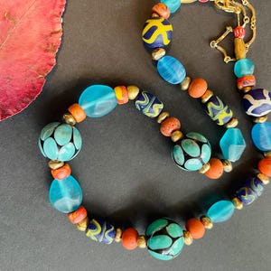 May include: A colorful necklace featuring a variety of beads in shades of blue, orange, and yellow. The necklace includes round, oval, and patterned beads, connected by gold-colored metal accents. The necklace is displayed on a dark surface.