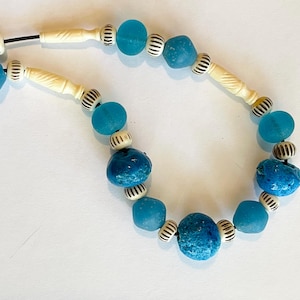 以下が含まれることがあります： 青いガラスビーズ、クリーム色の彫刻された骨のようなビーズ、白と黒のストライプのビーズを特徴とするネックレス。 ネックレスは白い表面に表示されています。