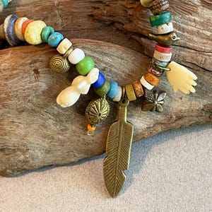 May include: A colorful beaded necklace with various charms, including a brass feather pendant, a small bird, and a hand-shaped charm. The beads are in shades of blue, green, yellow, and white. The necklace is displayed on a piece of driftwood.