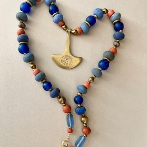 Peut inclure: Un collier artisanal avec un pendentif en laiton orné d'un motif en spirale. Le collier est composé de perles bleues, corail et dorées. Les perles sont de formes et de tailles variées, créant un design unique. Le collier est fermé par un fermoir doré.
