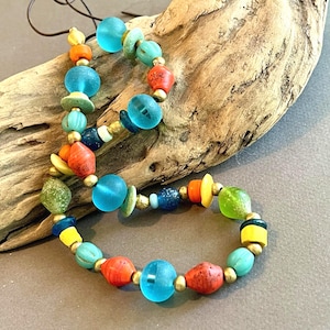 May include: A colorful beaded necklace with a variety of shapes and sizes. The beads are in shades of blue, orange, red, yellow, and green, with gold-colored spacers. The necklace is displayed on a piece of driftwood.