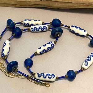 May include: A necklace featuring cylindrical white beads with blue leaf patterns, interspersed with round dark blue beads, small metallic spacers, and thin blue tube beads. The piece includes a gold-toned metal lobster clasp and an adjustable chain.