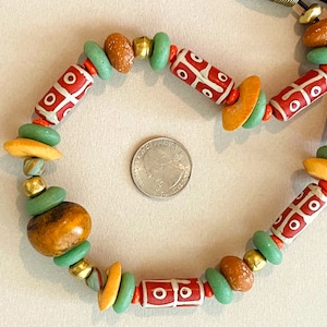 May include: A handcrafted necklace featuring a variety of beads in earthy tones. The necklace includes red and white patterned cylindrical beads, green disc beads, and orange and brown accent beads. The design is strung on a dark cord.