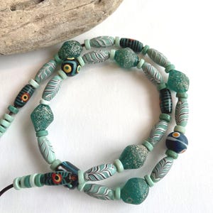 May include: A necklace with a selection of glass beads in teal, green, and white. The beads display diverse patterns, including leaf-like designs and speckled textures. The necklace is strung on a dark cord.