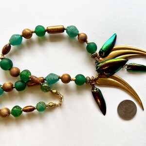 May include: A green and gold necklace with a large, ornate pendant featuring iridescent beetle wings and gold accents. The necklace is made with round glass beads and wooden beads.