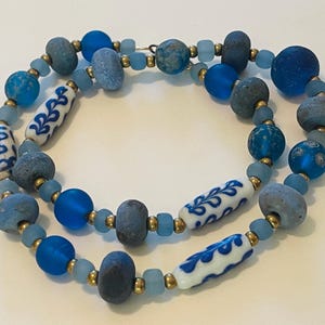 May include: A necklace featuring a mix of blue and grey matte beads, translucent light blue beads, and cylindrical white beads with a blue leaf pattern, all separated by small metallic gold-toned spacer beads arranged in a circular pattern.