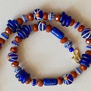 May include: A blue and brown glass bead necklace with a gold clasp. The beads are various shapes and sizes, including round, oval, and rectangular. The necklace is approximately 50 cm long.