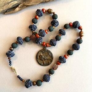 May include: A necklace made of denim, lava rock, and copper beads with a bronze pendant featuring a bird design.