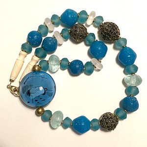 May include: A handcrafted necklace featuring a variety of beads in shades of blue, clear, and silver. The necklace includes round, textured, and irregular-shaped beads. A large, round blue pendant with a crane design is a focal point.