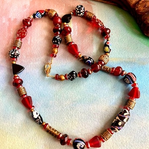 Peut inclure: Un collier artisanal composé de perles rouges, noires et dorées. Le collier comprend des perles rondes, en forme de goutte et cylindriques avec des motifs et des dessins complexes. Le collier est présenté sur une surface colorée avec un élément en bois en arrière-plan.