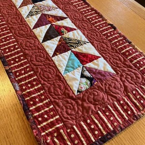 May include: A rectangular quilted table runner featuring a central column of multi-colored fabric triangles in various patterns, surrounded by a solid dusty rose border with swirl stitching and an outer edge of striped fabric with white dots.