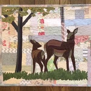May include: A rectangular fabric wall hanging featuring a quilted patchwork background of various floral and striped patterns. The scene depicts a dark brown deer and a smaller fawn standing in green fabric grass beneath a dark tree with green leaves.