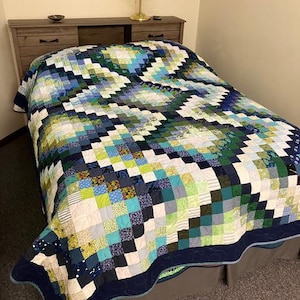May include: A handmade quilt with a patchwork design in shades of blue, green, and white. The quilt features a dark blue border with a decorative stitch. The bed is covered with the quilt, showcasing its intricate pattern and craftsmanship.