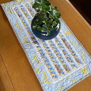 May include: A handmade, quilted table runner with a blue, yellow, and white geometric pattern. A dark blue ceramic pot holds a green plant. The runner is placed on a wooden table, creating a decorative and functional home accent.