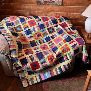 May include: A colorful patchwork quilt with a square pattern, featuring various fabric squares in shades of blue, red, yellow, and green. The quilt is draped over a couch, showcasing its intricate design and craftsmanship. The quilt's border is a mix of colors.