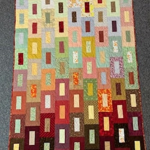 May include: A handmade quilt with a patchwork design. The quilt features a variety of rectangular blocks in shades of red, brown, yellow, green, and orange. The blocks are arranged in a grid pattern, creating a visually appealing and cozy textile.