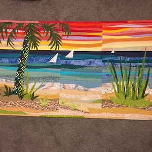 May include: A colorful fabric art piece depicting a beach scene. The artwork features a palm tree, sailboats, and tall grass against a sunset sky. The ocean is represented with various shades of blue and green.