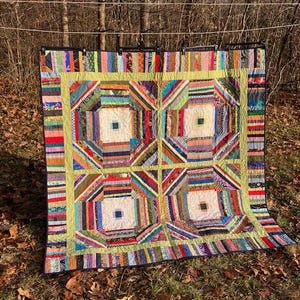 May include: A handmade quilt with a patchwork design. The quilt features a central pattern of four large squares with a light-colored center, surrounded by colorful, geometric shapes. The border is a bright green, and the quilt is displayed outdoors.