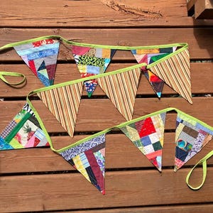 May include: A collection of colorful, handmade fabric bunting flags. Each triangular flag features a patchwork design with various patterns and colors, including stripes, floral prints, and geometric shapes. The flags are strung together with a green ribbon.