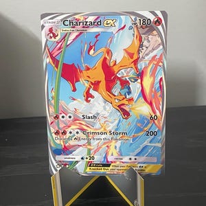 Puede incluir: Una carta coleccionable Charizard EX, con un dragón naranja llameante sobre un fondo azul y rojo. La carta muestra los nombres de ataque "Slash" y "Crimson Storm", con 180 PV. La carta se muestra en un soporte.