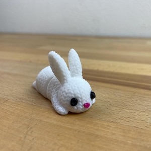 Könnte beinhalten: Eine weiße, strukturierte Hasenfigur mit schwarzen Augen und einer rosa Nase. Der Hase liegt auf einer Holzoberfläche. Die Figur hat lange Ohren und ein detailliertes, gestricktes Aussehen.