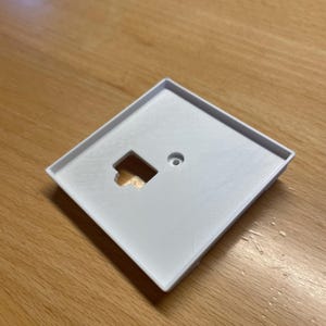 May include: A white, square light switch cover with a rectangular opening and a small circular hole. The cover is made of a smooth, matte material and sits on a wooden surface. The design is simple and functional.