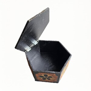Könnte beinhalten: Eine schwarze, sechseckige Holzbox mit leicht geöffnetem Deckel. Die Box hat ein dekoratives Blumenmuster in Orange und Gold an den Seiten. Der Innenraum der Box ist ebenfalls schwarz.
