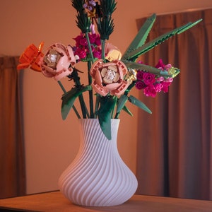 May include: A white, swirled vase holds a colorful bouquet of flowers. The flowers are made of plastic and include orange, pink, and purple blooms with green stems and leaves. The vase sits on a wooden table.