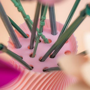 May include: A close-up of a pink floral arrangement holder with green and gray stems. The holder has a ridged, pleated design on the base and a perforated top to hold the stems in place. The background is a soft peach color.