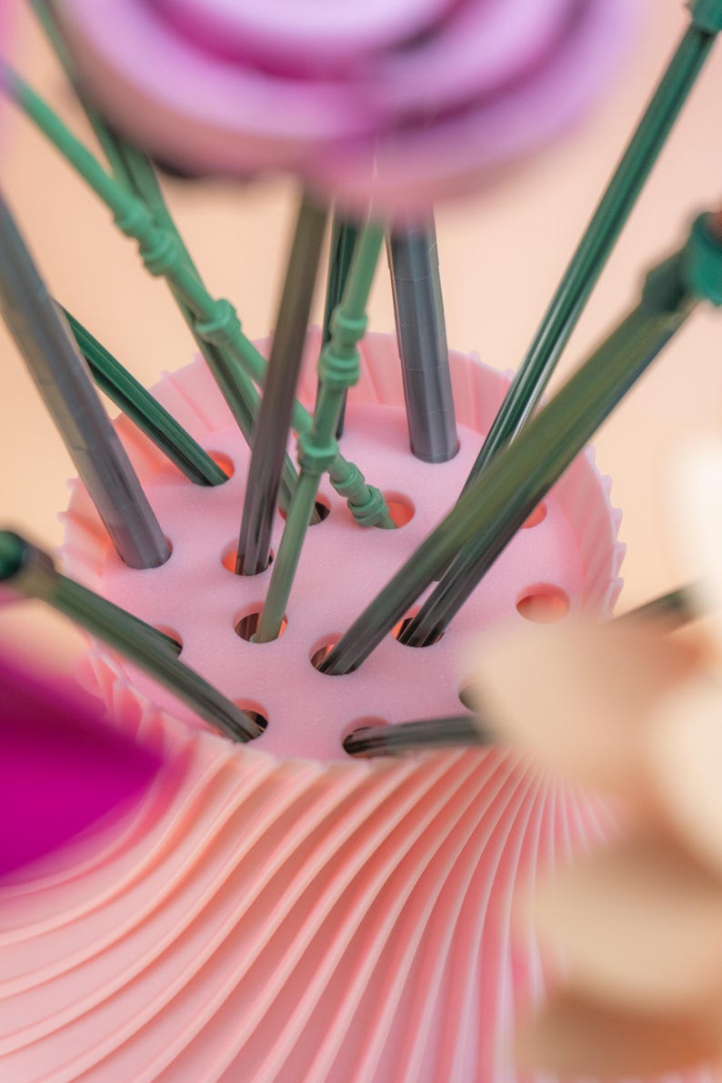 May include: A close-up of a pink floral arrangement holder with green and gray stems. The holder has a ridged, pleated design on the base and a perforated top to hold the stems in place. The background is a soft peach color.