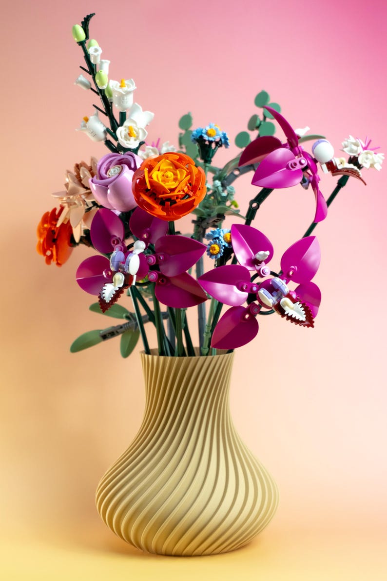 May include: A vibrant bouquet of colorful flowers in a gold vase. The arrangement includes orange, purple, and white blooms, with green stems and leaves. The vase has a textured, swirling design.