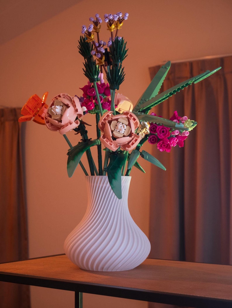 May include: A white, swirled vase holds a colorful bouquet of flowers. The flowers are made of plastic and include orange, pink, and purple blooms with green stems and leaves. The vase sits on a wooden table.